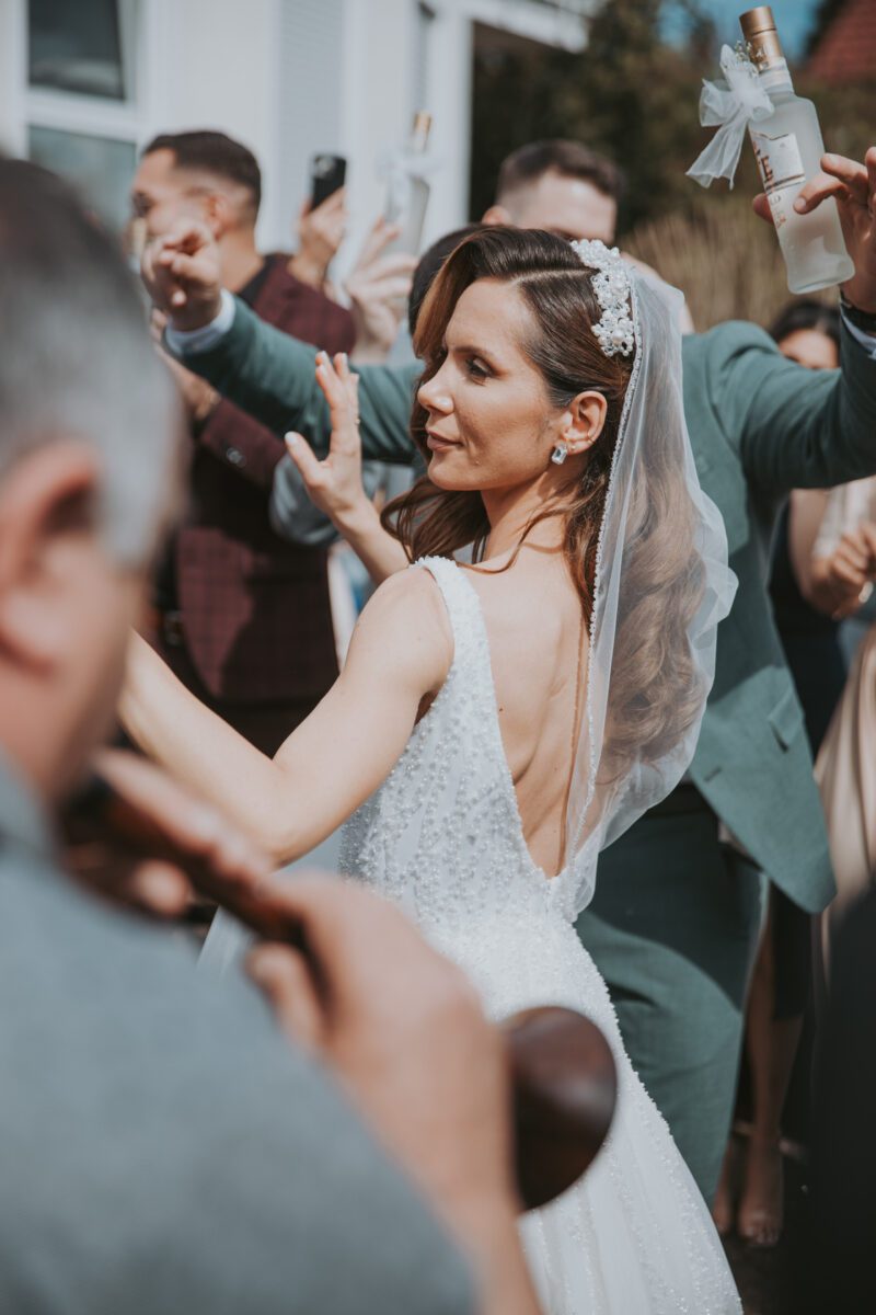 Eine Braut in einem weißen, ärmellosen, rückenfreien Kleid und Schleier tanzt vor einem Gebäude, umgeben von Gästen, die Flaschen halten und ihre Hände heben. Ihr langes, welliges Haar fällt ihr über den Rücken, geschmückt mit einem dekorativen Haarteil. Im Vordergrund ist teilweise ein Musiker zu sehen, der ein Instrument spielt und den Moment durch die Linse von Hochzeitsfotografie festhält.
 - Elis Wedding - Hochzeitsfotograf Frankfurt