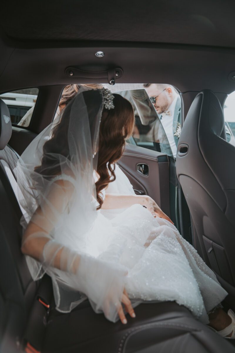 Eine Braut in weißem Kleid und Schleier sitzt auf dem Rücksitz eines Autos und rafft vorsichtig ihr Kleid. Ihr Haar ist in weiche Locken gestylt und mit einem funkelnden Haarteil geschmückt. Durch die offene Tür bietet ihr während eines Fotoshootings ein Mann in hellem Anzug und Sonnenbrille seine Hand an, um ihr herauszuhelfen. - Elis Wedding - Hochzeitsfotograf Frankfurt