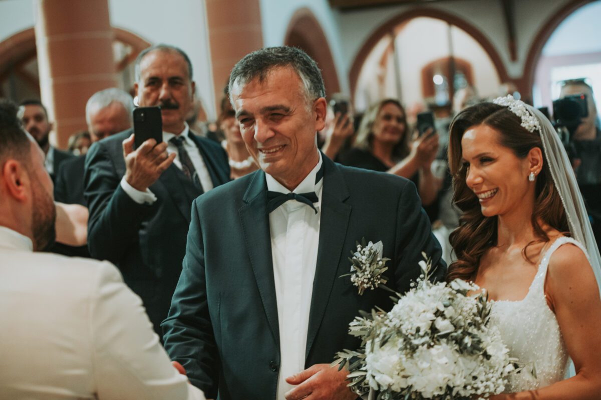 Eine Braut in einem weißen Kleid und einem Blumenkopfschmuck lächelt und hält einen Strauß weißer Blumen in der Hand. Sie steht neben einem älteren Mann in einem dunklen Anzug mit einer Ansteckblume. Der Mann schüttelt während des Fotoshootings einem anderen Mann in einem weißen Anzug die Hand. Im Hintergrund sind Gäste in Abendgarderobe zu sehen, einige machen Fotos. - Elis Wedding - Hochzeitsfotograf Frankfurt