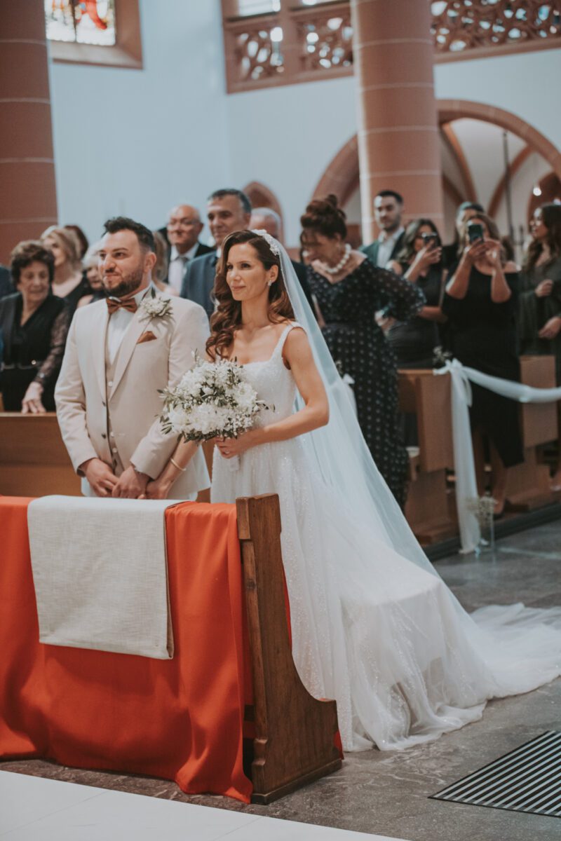 Braut und Bräutigam stehen lächelnd am Altar einer Kirche. Die Braut trägt ein weißes Brautkleid mit langem Schleier und hält einen Strauß weißer Blumen in der Hand. Der Bräutigam trägt einen hellbeigen Anzug. Hinter ihnen sitzen Gäste, einige stehen und machen ein Fotoshooting. Die Kirche hat hohe, rote Säulen und Bogenfenster. - Elis Wedding - Hochzeitsfotograf Frankfurt