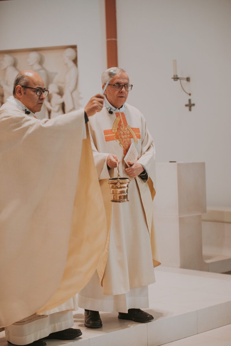 Zwei Geistliche führen in einer Kirche eine religiöse Zeremonie durch. Sie sind in cremefarbene Gewänder gekleidet. Einer hält ein Weihrauchfass, aus dem Rauch aufsteigt. Im Hintergrund sind ein Kreuz und religiöse Reliefs zu sehen, deren feierlicher Ausdruck die Bedeutung des Rituals unterstreicht, das einer standesamtlichen Trauung ähnelt. - Elis Wedding - Hochzeitsfotograf Frankfurt