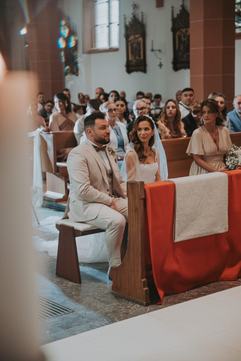 Braut und Bräutigam sitzen vor einer Kirche, mit Blick auf den Altar. Der Bräutigam trägt einen hellen Anzug und die Braut ein weißes Kleid. Sie sind von Gästen umgeben, die in Kirchenbänken unter Buntglasfenstern und religiösen Kunstwerken sitzen. Die Szene wurde von einem Hochzeitsfotografen aufgenommen und ist sanft beleuchtet, was eine warme Atmosphäre schafft. - Elis Wedding - Hochzeitsfotograf Frankfurt