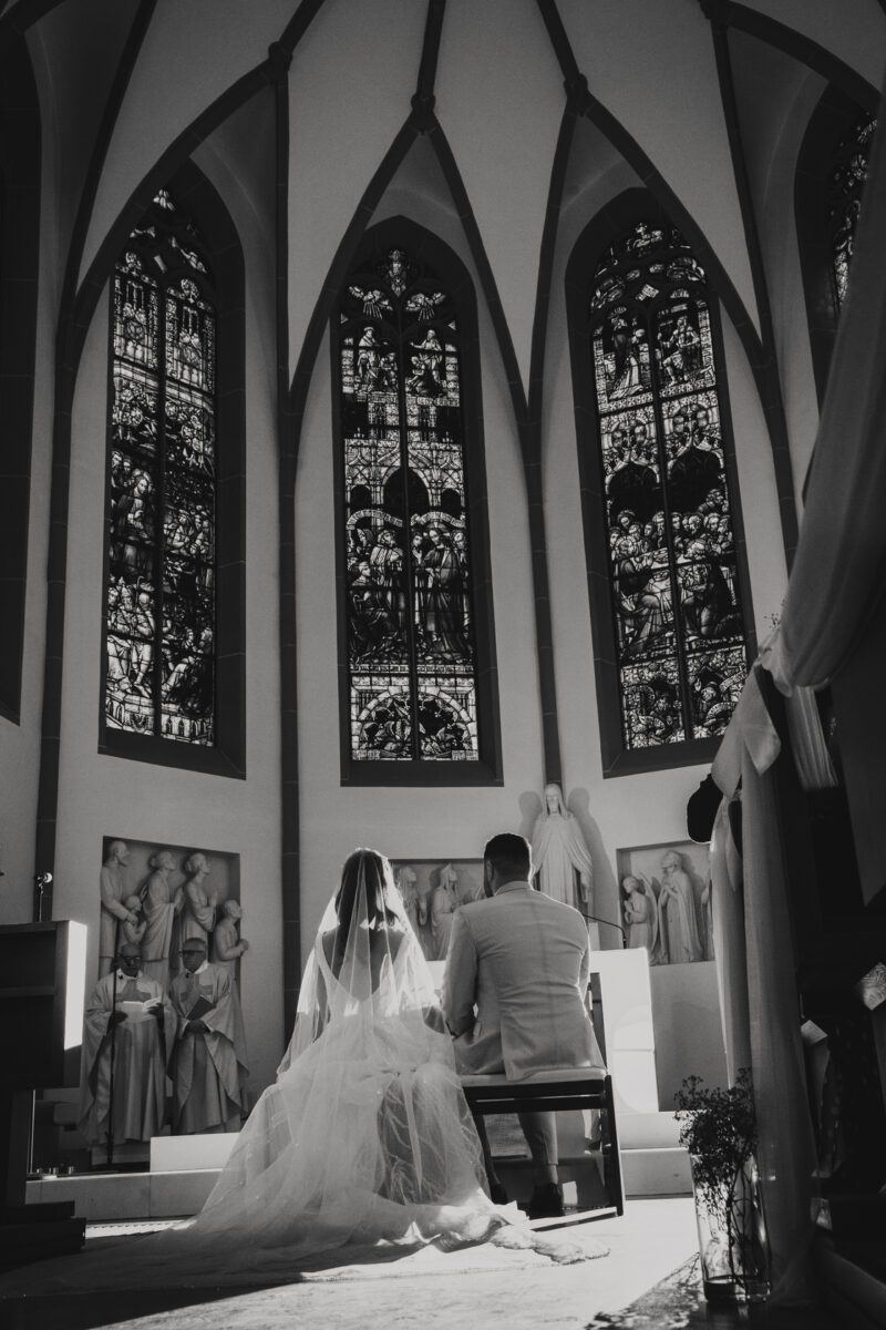 Eine Schwarz-Weiß-Fotografie fängt die standesamtliche Trauung eines Paares in einer Kirche in Frankfurt ein. Das Paar sitzt mit dem Rücken zur Kamera am Altar, der mit detailreichen Statuen und drei großen, kunstvollen Buntglasfenstern geschmückt ist. Die Braut trägt einen langen Schleier und ein langes Kleid, und der Bräutigam trägt einen hellen Anzug. Natürliches Licht erhellt die Szene. - Elis Wedding - Hochzeitsfotograf Frankfurt