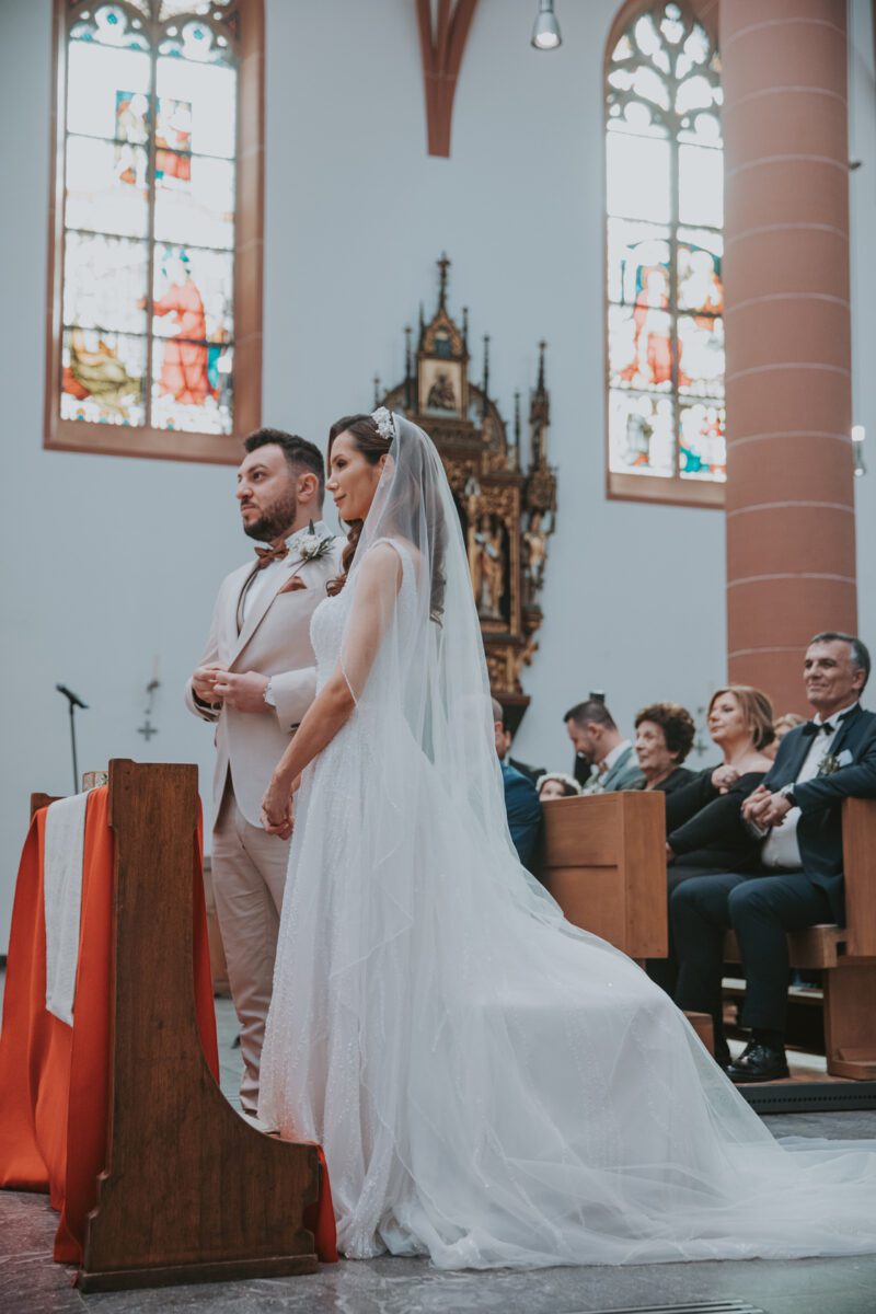 Braut und Bräutigam stehen in einer Kirche am Altar, perfekt eingefangen auf diesem Foto. Die Braut trägt ein weißes Spitzenkleid und einen Schleier, während der Bräutigam einen hellen Anzug trägt. Sie blicken nach vorne und haben die Hände gefaltet. Hinter ihnen sitzen Hochzeitsgäste in Kirchenbänken mit Buntglasfenstern und religiösem Dekor im Hintergrund. - Elis Wedding - Hochzeitsfotograf Frankfurt