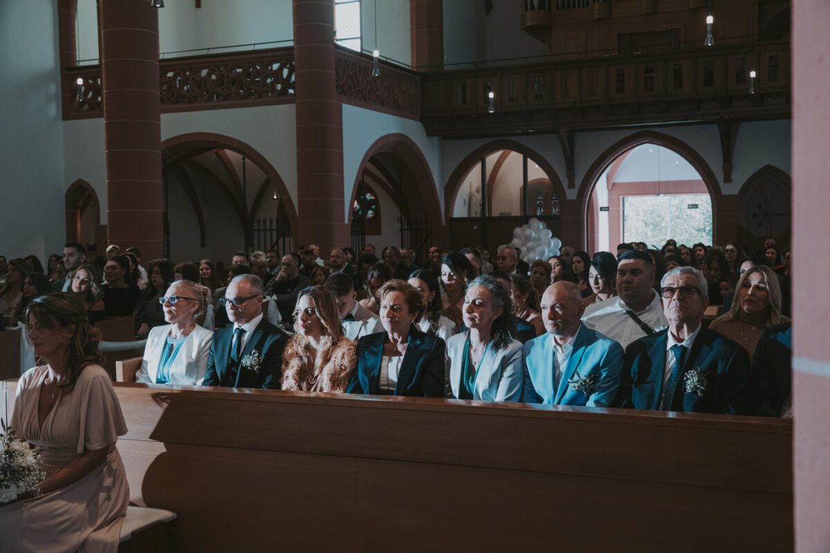 Eine Gruppe gut gekleideter Menschen sitzt während einer Zeremonie aufmerksam in den Holzbänken einer wunderschön geschmückten Kirche in Frankfurt. Das Publikum, bestehend aus Männern und Frauen, wird von natürlichem Licht erhellt, das durch die hohen Bogenfenster strömt, als ob es sich auf ein Fotoshooting vorbereitet hätte, und hebt die architektonischen Details der Kirche hervor. - Elis Wedding - Hochzeitsfotograf Frankfurt