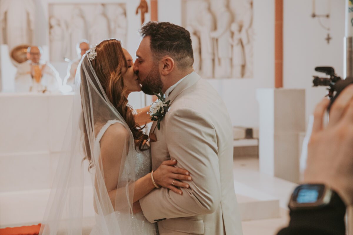 Ein frisch vermähltes Paar küsst sich während seiner standesamtlichen Trauung drinnen am Altar. Die Braut, die ein weißes Kleid und einen Schleier trägt, hält den Bräutigam, der einen hellen Anzug trägt. Im Hintergrund ist ein Priester zu sehen, während jemand rechts den Moment mit einer Kamera festhält und diese wunderschöne Fotografie verewigt. - Elis Wedding - Hochzeitsfotograf Frankfurt