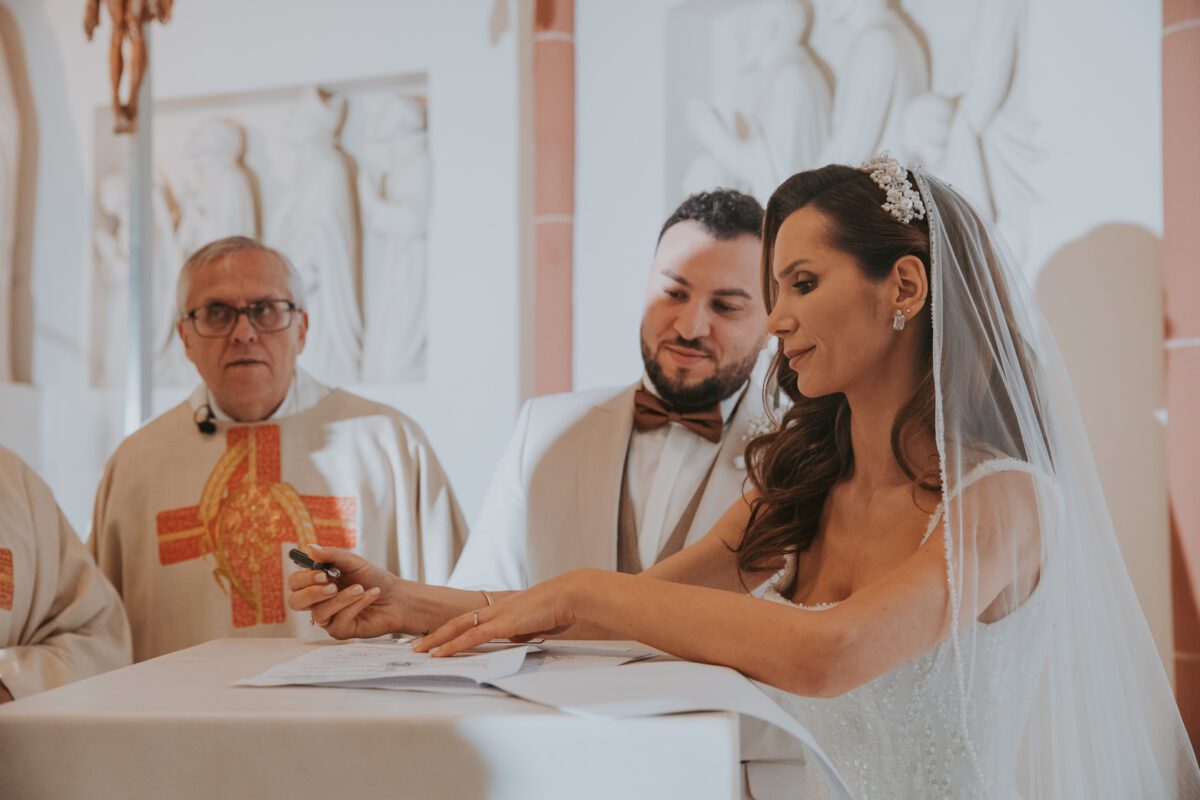 Eine Braut in einem weißen Brautkleid und Schleier unterschreibt ein Dokument auf einem Tisch, den der Bräutigam in einem beigen Anzug und einer braunen Fliege hält. Zwei Geistliche in weißen Gewändern mit roten und goldenen Kreuzen stehen hinter ihnen, während der Hochzeitsfotograf diesen kostbaren Moment in der Kirche mit weißen Wänden und religiösen Reliefs festhält. - Elis Wedding - Hochzeitsfotograf Frankfurt