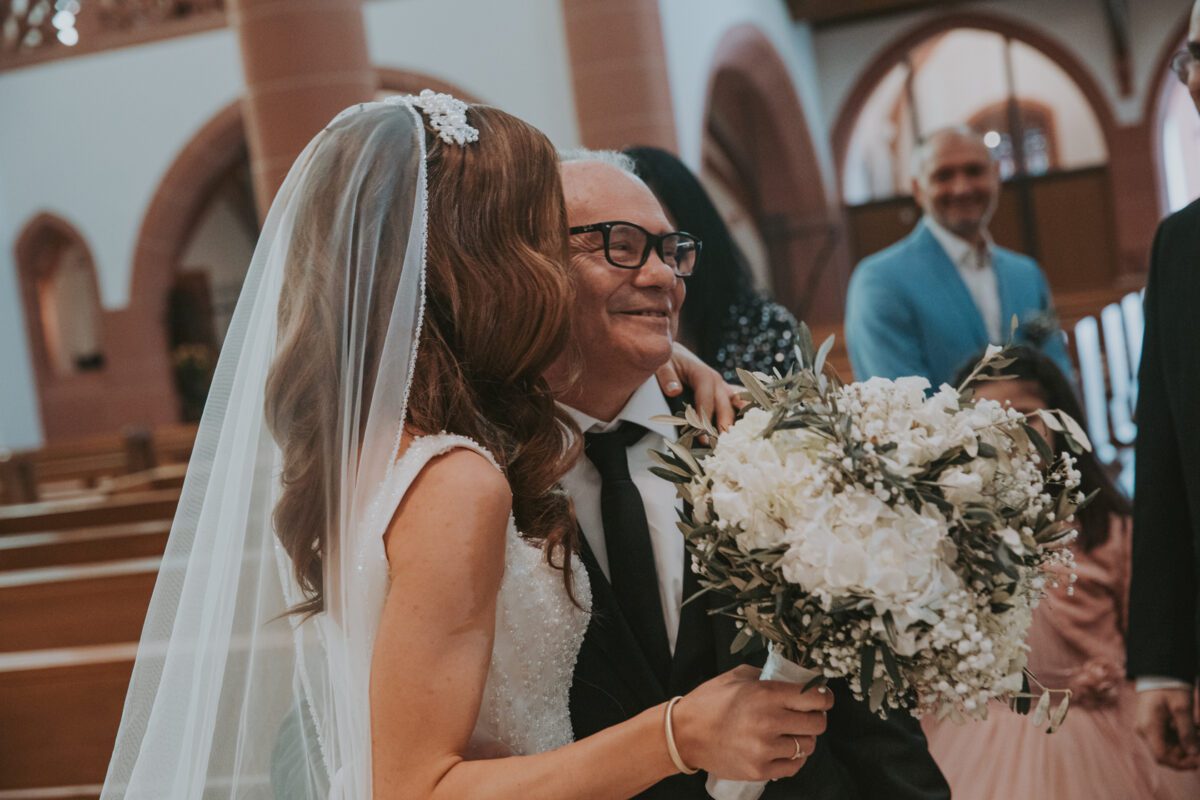Eine Braut in weißem Kleid und Schleier, die einen Strauß weißer Blumen hält, steht lächelnd neben einem älteren Mann im dunklen Anzug mit Brille. Die Kulisse erinnert an eine standesamtliche Trauung mit Holzbänken und Bogenfenstern. Im Hintergrund sind ein Mann im hellblauen Anzug und ein Mädchen im rosa Kleid zu sehen. - Elis Wedding - Hochzeitsfotograf Frankfurt