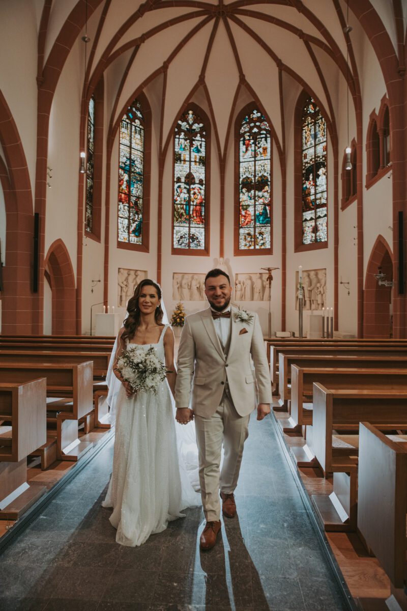 Braut und Bräutigam gehen während ihrer standesamtlichen Trauung den Mittelgang einer Kirche mit hohen Decken und wunderschönen Buntglasfenstern entlang. Die Braut trägt ein langes weißes Kleid und hält einen Blumenstrauß, während der Bräutigam einen hellen Anzug trägt. Die Kirchenbänke sind leer und Licht strömt durch die Fenster dahinter herein. - Elis Wedding - Hochzeitsfotograf Frankfurt
