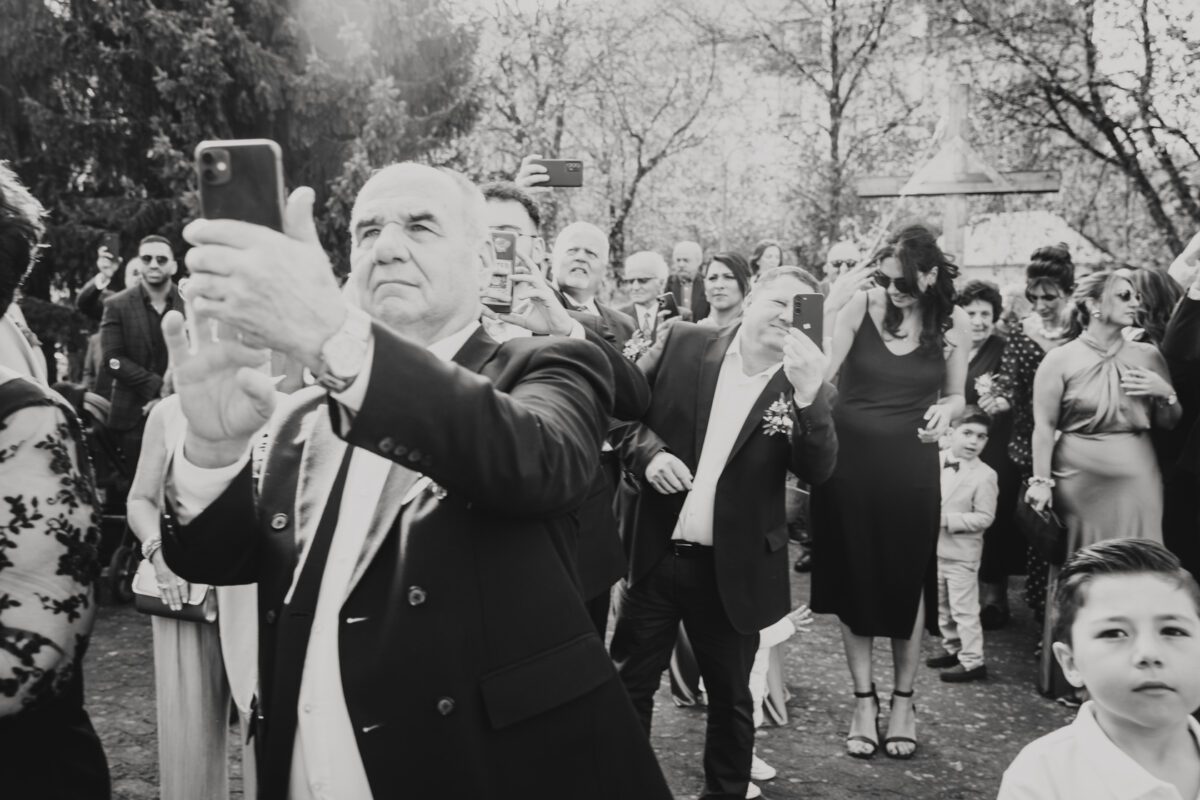 Ein Schwarzweißfoto einer Versammlung im Freien in Frankfurt. Die Leute halten ihre Telefone hoch und scheinen einen Moment festzuhalten. Ein älterer Mann im Vordergrund trägt einen Anzug und gestikuliert, als würde er ein Foto machen. Andere Teilnehmer, darunter Frauen, Kinder und Männer in Abendgarderobe, unterhalten sich zwischen Bäumen im Hintergrund mit dem Hochzeitsfotografen. - Elis Wedding - Hochzeitsfotograf Frankfurt