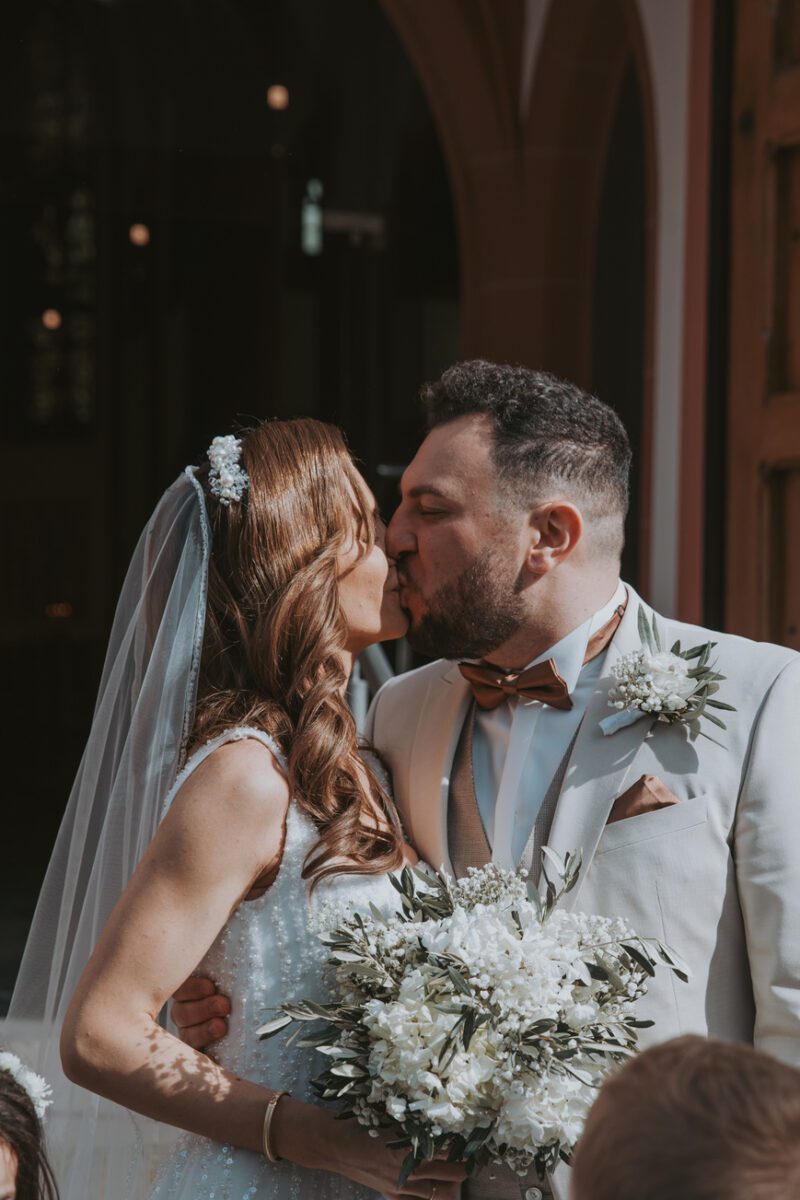 Braut und Bräutigam, die sich beim Kennenlernen kennengelernt haben, küssen sich an ihrem Hochzeitstag. Die Braut in einem weißen Kleid und Schleier hält einen Strauß weißer Blumen in der Hand. Der Bräutigam, der einen hellen Anzug mit einer braunen Fliege und einer Blumenboutonniere trägt, hat seinen Arm um sie gelegt, während ihr Hochzeitsfotograf den Moment vor gewölbten Türen festhält. - Elis Wedding - Hochzeitsfotograf Frankfurt