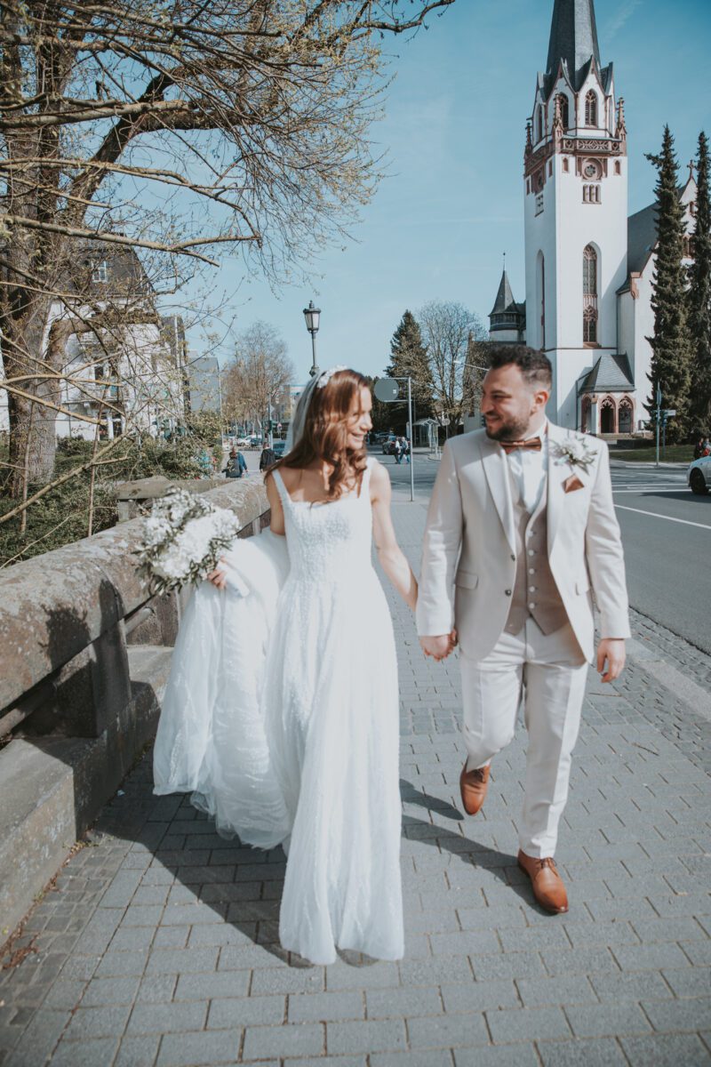 Eine Braut in einem weißen Brautkleid hält ihr Kleid, während sie Hand in Hand mit einem Bräutigam in einem beigen Anzug geht, aufgenommen von ihrem Hochzeitsfotografen während eines Fotoshootings in Frankfurt. Sie schlendern einen Steinweg in der Nähe der Straße entlang, hinter ihnen eine große Kirche. Beide lächeln und schauen sich zwischen spärlichen Bäumen unter einem klaren Himmel an. - Elis Wedding - Hochzeitsfotograf Frankfurt