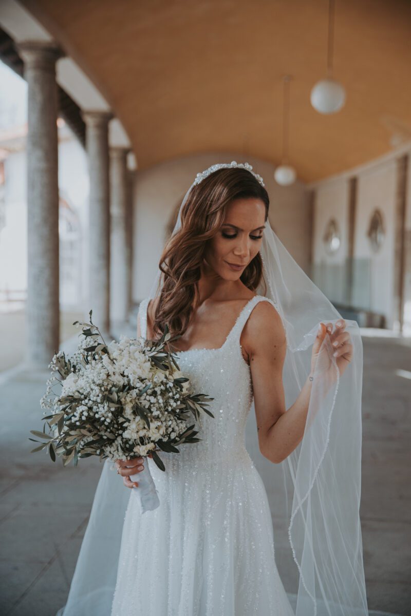Eine Braut in einem weißen, ärmellosen Kleid, das mit zarten Perlen und Stickereien geschmückt ist, posiert anmutig. In einer Hand hält sie einen weißen Blumenstrauß und hebt mit der anderen sanft ihren langen, durchsichtigen Schleier. Die elegante Szene, die von einem Hochzeitsfotografen in Frankfurt festgehalten wurde, zeigt eine gewölbte bernsteinfarbene Decke und hängende Kugellampen im Hintergrund. - Elis Wedding - Hochzeitsfotograf Frankfurt