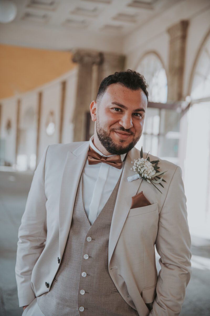 Ein Mann in einem cremefarbenen Anzug mit brauner Fliege und Einstecktuch lächelt, während er während eines Fotoshootings in Frankfurt drinnen steht. Er hat kurze dunkle Haare und einen Bart und trägt eine Ansteckblume am linken Revers. Der Hintergrund umfasst Bogenfenster, hohe Säulen und sanftes Tageslicht. - Elis Wedding - Hochzeitsfotograf Frankfurt