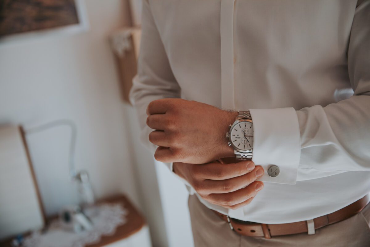 Eine Person in weißem Hemd und beiger Hose rückt eine silberne Armbanduhr an ihrem linken Handgelenk zurecht. Das Foto, aufgenommen von einem Hochzeitsfotografen in Frankfurt, wurde in einem Innenbereich mit sanfter Beleuchtung und einem unscharfen Hintergrund aufgenommen, der einen Teil eines Bettes und einen Nachttisch mit einem Spitzendeckchen zeigt. - Elis Wedding - Hochzeitsfotograf Frankfurt