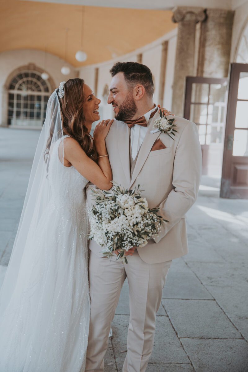 Braut und Bräutigam stehen dicht beieinander, lächeln und schauen sich in die Augen. Die Braut trägt ein weißes Kleid und einen Schleier und hält einen Strauß weißer Blumen in der Hand. Der Bräutigam trägt einen hellbeigen Anzug mit passender Boutonnière und Einstecktuch. Sie befinden sich in einem Saal mit Steinsäulen und Bogenfenstern, perfekt für unvergessliche Fotos. - Elis Wedding - Hochzeitsfotograf Frankfurt