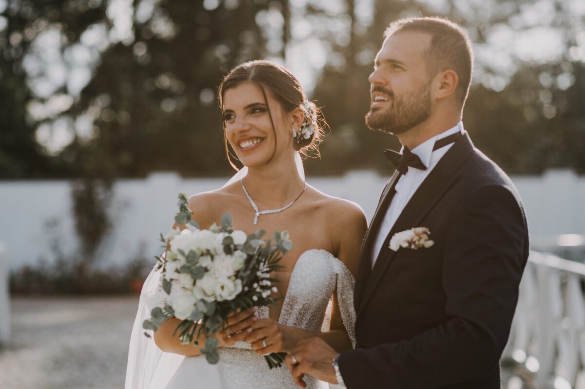 Eine Braut in einem trägerlosen weißen Brautkleid und ein Bräutigam in einem schwarzen Smoking stehen draußen, lächeln und blicken nach rechts. Die Braut hält einen Strauß weißer Blumen, während das Sonnenlicht durch die Bäume hinter ihnen fällt und eine warme und fröhliche Atmosphäre während ihres besonderen Fotoshootings durch ihren Hochzeitsfotografen in Frankfurt schafft. - Elis Wedding - Hochzeitsfotograf Frankfurt