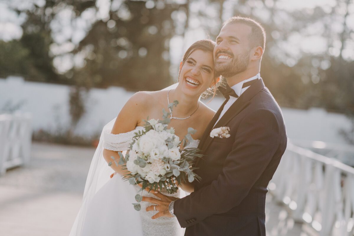 Eine fröhliche Braut und ein Bräutigam stehen Seite an Seite im Freien und lächeln strahlend während ihres Fotoshootings. Die Braut trägt ein weißes schulterfreies Brautkleid und hält einen Strauß weißer Blumen und Grünpflanzen in der Hand. Der Bräutigam trägt einen schwarzen Smoking mit Fliege. Sie scheinen auf einer Brücke in Frankfurt zu stehen, während sanftes Sonnenlicht durch die Bäume fällt. - Elis Wedding - Hochzeitsfotograf Frankfurt