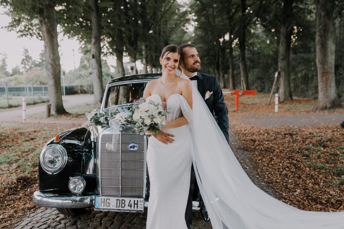 Eine Braut in einem trägerlosen weißen Kleid und ein Bräutigam in einem schwarzen Anzug posieren für ein Fotoshooting vor einem schwarzen Oldtimer. Die Braut hält einen Strauß weißer Blumen und Grünpflanzen, hinter ihr weht ein langer Schleier. Sie sind umgeben von hohen Bäumen und einem mit Laub bedeckten Weg, perfekt eingefangen von ihrem Hochzeitsfotografen. - Elis Wedding - Hochzeitsfotograf Frankfurt