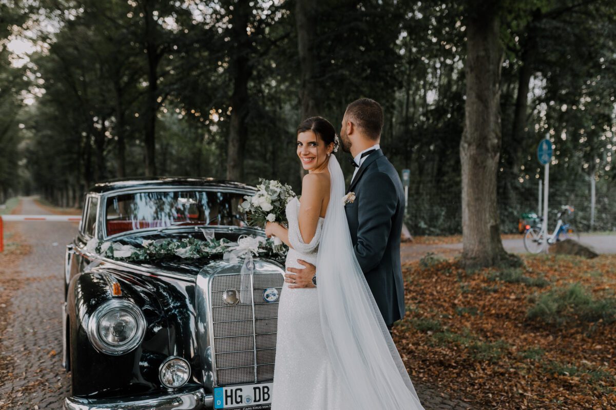 Braut und Bräutigam stehen neben einem mit Blumen geschmückten schwarzen Oldtimer. Die Braut, in einem weißen Kleid mit langem Schleier, blickt zurück und lächelt, während sie einen Blumenstrauß hält. Der Bräutigam, in einem dunklen Anzug, steht mit dem Rücken zur Kamera und blickt die Braut an. Ein Waldstück bietet einen ruhigen Hintergrund – ein perfekter Moment, eingefangen von ihrem Hochzeitsfotografen. - Elis Wedding - Hochzeitsfotograf Frankfurt