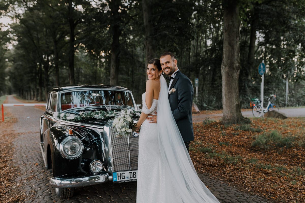 Ein frisch vermähltes Paar in Abendgarderobe posiert neben einem mit Blumen geschmückten schwarzen Oldtimer, aufgenommen während ihres Fotoshootings. Die Braut trägt ein weißes Kleid mit langer Schleppe und hält einen Blumenstrauß, während der Bräutigam einen Smoking trägt. Sie stehen auf einem von Bäumen umgebenen Kopfsteinpflasterweg, im Hintergrund sind eine rot-weiße Absperrung und ein Fahrrad zu sehen. - Elis Wedding - Hochzeitsfotograf Frankfurt