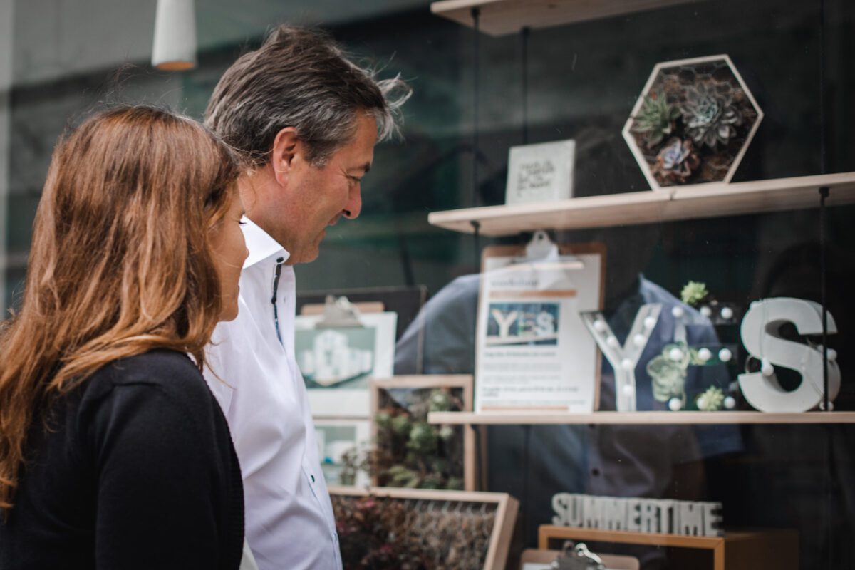 Ein Mann und eine Frau stehen eng beieinander und schauen in ein Schaufenster. Im Schaufenster sind verschiedene Dekorationsgegenstände zu sehen, darunter Topfpflanzen, gerahmte Kunstwerke sowie die Buchstaben „Y“ und „S“. Beide Personen wirken interessiert und aufmerksam auf die Auslage – vielleicht eine ideale Kulisse für einen Hochzeitsfotografen in Frankfurt. - Elis Wedding - Hochzeitsfotograf Frankfurt