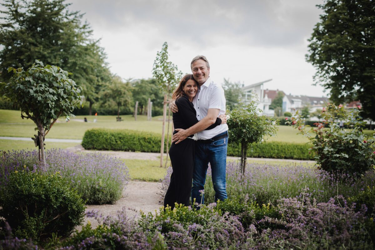 Ein Paar umarmt sich inmitten eines üppigen Gartens, umgeben von blühendem Lavendel und grünen Büschen. Beide lächeln, die Frau trägt ein schwarzes Kleid und der Mann ein weißes Hemd und blaue Jeans. Hinter ihnen fängt ein Hochzeitsfotograf jedes Detail ein, während sich der Park mit Bäumen und ordentlich geschnittenen Hecken in die Ferne erstreckt. - Elis Wedding - Hochzeitsfotograf Frankfurt