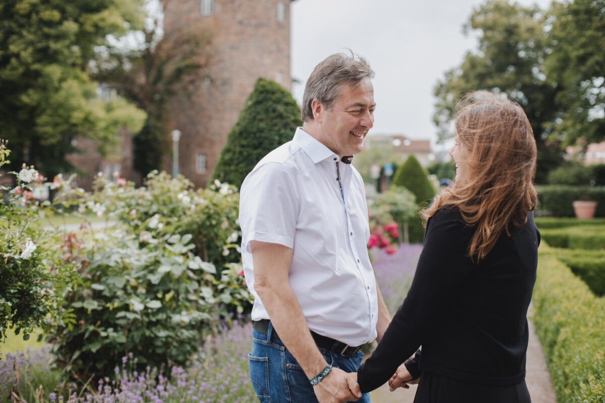 Ein Mann und eine Frau lächeln und halten sich an den Händen in einem Garten mit bunten Blumen und grünen Sträuchern. Der Mann in weißem Hemd und Jeans steht der Frau gegenüber, die lange Haare hat und ein schwarzes Outfit trägt. Mit einem Steinturm und Bäumen im Hintergrund könnte dies der perfekte Ort für einen Hochzeitsfotografen in Frankfurt sein. - Elis Wedding - Hochzeitsfotograf Frankfurt