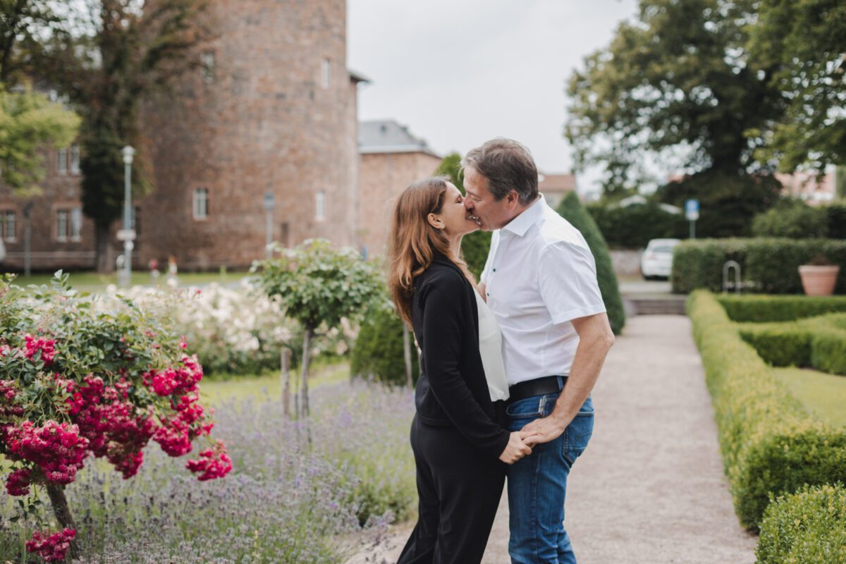 Ein Paar steht lachend und umarmend in einem Park. Der Mann in weißem Hemd und Jeans lehnt sich eng an die Frau, die ein schwarzes Outfit trägt. Sie sind umgeben von üppigem Grün, leuchtend rosa und lila Blumen und einem Steinweg. Im Hintergrund ist ein historisches Steingebäude zu sehen – eine perfekte Szene für eine Fotografie. - Elis Wedding - Hochzeitsfotograf Frankfurt