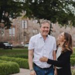 Ein Mann und eine Frau stehen draußen vor einem großen, historischen Backsteingebäude mit hohen Fenstern in Frankfurt. Der Mann trägt ein weißes Hemd und Jeans und lächelt, während die Frau, die einen dunklen Blazer über einem weißen Oberteil trägt, lachend neben ihm steht. Sie sind von ordentlich geschnittenen Hecken und Grün umgeben – ein perfekter Moment für eine Fotografie. - Elis Wedding - Hochzeitsfotograf Frankfurt