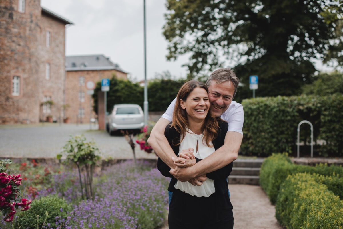 Eine Frau und ein Mann posieren draußen in einem Garten mit einem historischen Gebäude im Hintergrund. Der Mann umarmt die Frau spielerisch von hinten und beide lächeln freudig. Während ihres Frankfurter Fotoshootings blühen Lavendel und andere Blumen entlang eines von Grün gesäumten Kieswegs. - Elis Wedding - Hochzeitsfotograf Frankfurt