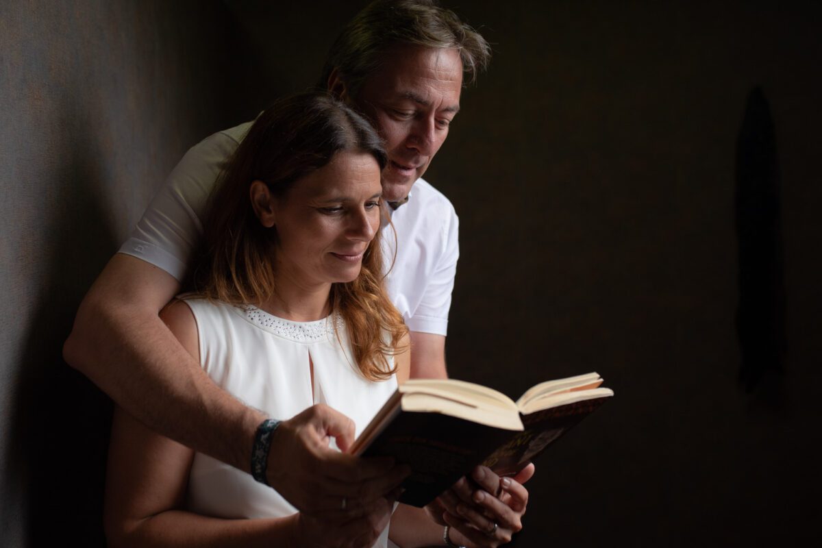 Ein Paar genießt einen ruhigen Moment des Kennenlernens in einem schwach beleuchteten Raum. Die Frau in einem weißen ärmellosen Oberteil hält ein offenes Buch, während der Mann in einem weißen Hemd hinter ihr steht und ihr über die Schulter blickt. Beide lächeln sanft. Es ist, als ob dieses zärtliche Fotoshooting ihre gemeinsame Intimität vor dem dunklen und verschwommenen Hintergrund einfängt. - Elis Wedding - Hochzeitsfotograf Frankfurt