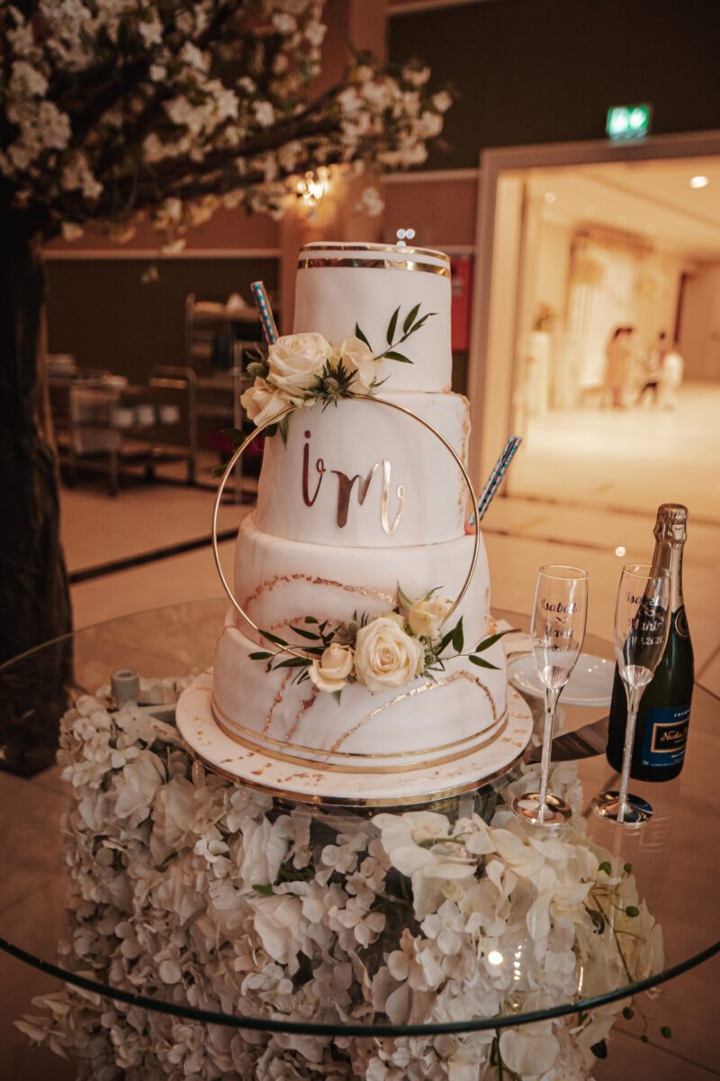 Eine dreistöckige Hochzeitstorte mit weißem Zuckerguss, goldenen Akzenten und frischen weißen Blumen wird auf einem Glastisch präsentiert. Die Initialen „V“ und „M“ sind auf der mittleren Etage deutlich zu sehen. Neben der Torte, perfekt für Ihr standesamtliches Trauungs-Fotoshooting, stehen eine Flasche Sekt und zwei Champagnergläser. - Elis Wedding - Hochzeitsfotograf Frankfurt