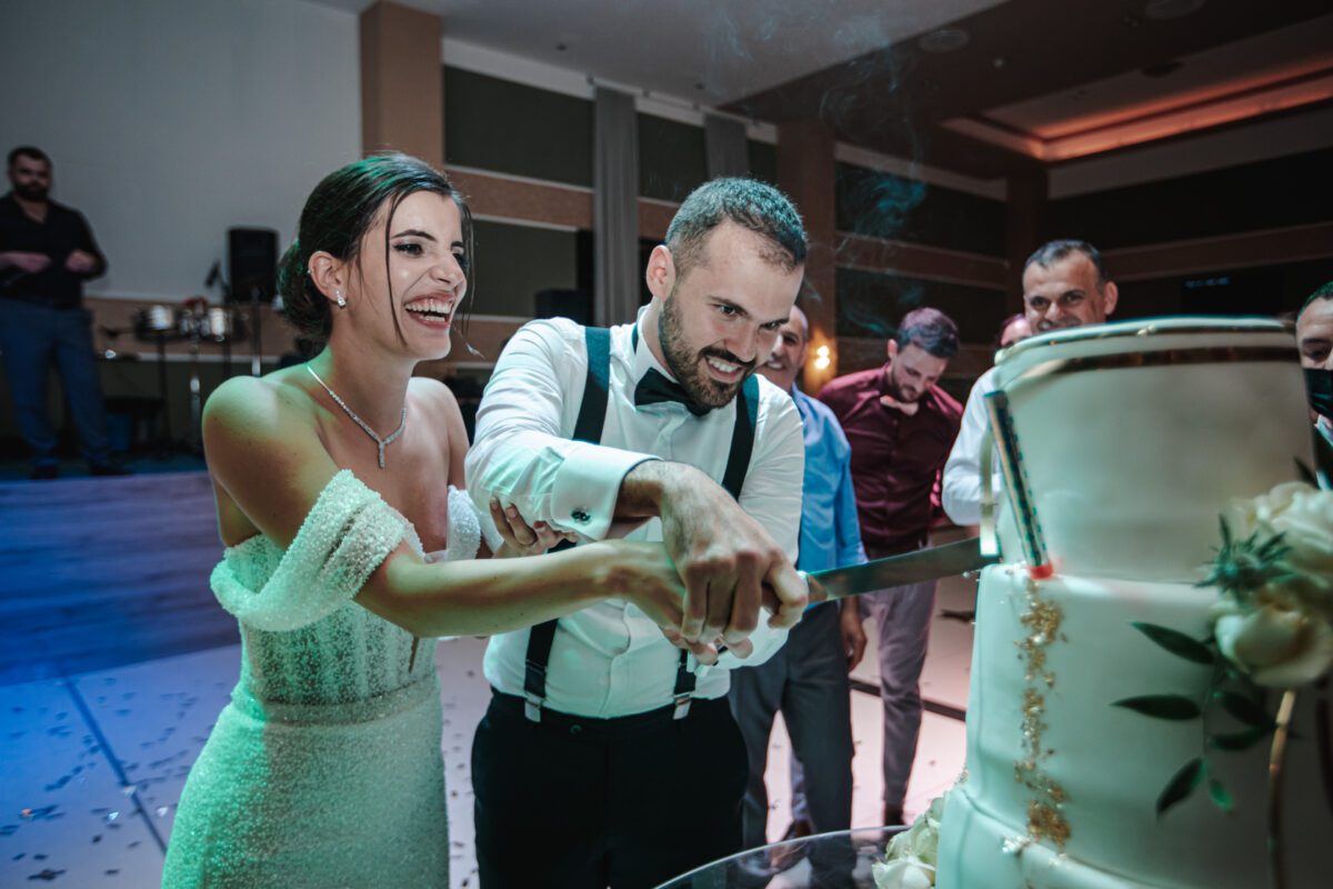Braut und Bräutigam schneiden während ihres Fotoshootings gemeinsam freudig eine dreistöckige Hochzeitstorte an. Die Braut in einem schulterfreien weißen Kleid lächelt strahlend, während der Bräutigam in einem weißen Hemd, schwarzen Hosenträgern und einer Fliege konzentriert bei der Sache ist. Die Gäste im Hintergrund beobachten und feiern den Moment, den ihr Hochzeitsfotograf festgehalten hat. - Elis Wedding - Hochzeitsfotograf Frankfurt