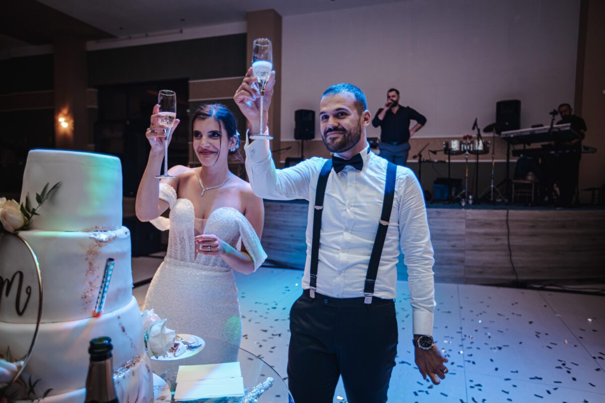 Braut und Bräutigam stehen zusammen und stoßen mit einem Champagnerglas an. Die Braut trägt ein weißes, schulterfreies Brautkleid und der Bräutigam ein weißes Hemd mit schwarzen Hosenträgern und Fliege. Im Hintergrund spielt eine Band auf einer Bühne in Frankfurt. Vor ihnen steht eine mehrstöckige Hochzeitstorte auf dem Tisch. - Elis Wedding - Hochzeitsfotograf Frankfurt
