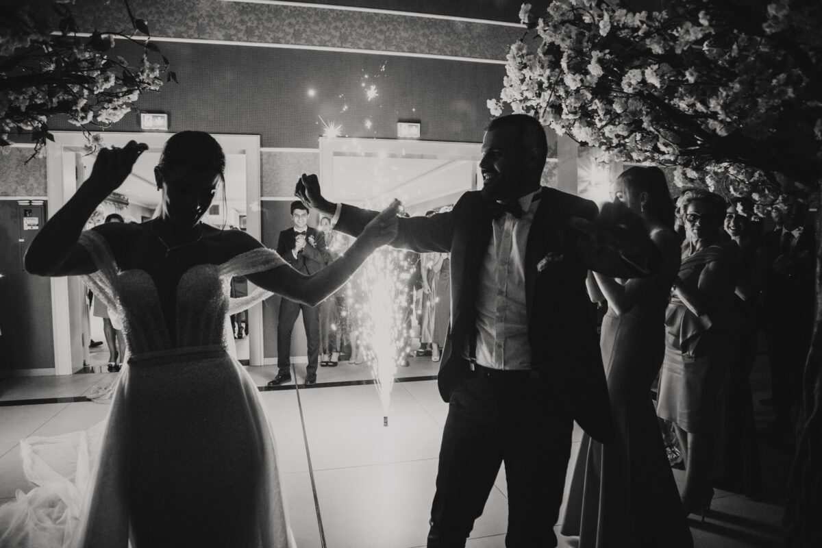 Ein Schwarzweißfoto fängt einen freudigen Moment bei einer Hochzeitsfeier ein. Die Frischvermählten tanzen Händchen haltend auf der Tanzfläche, umgeben von Gästen. Ein Hochzeitsfotograf fotografiert, während funkelnde Feuerwerke die Szene erhellen, während mit Blumen geschmückte Bäume das Paar umrahmen. Gäste in Abendgarderobe schauen glücklich zu. - Elis Wedding - Hochzeitsfotograf Frankfurt