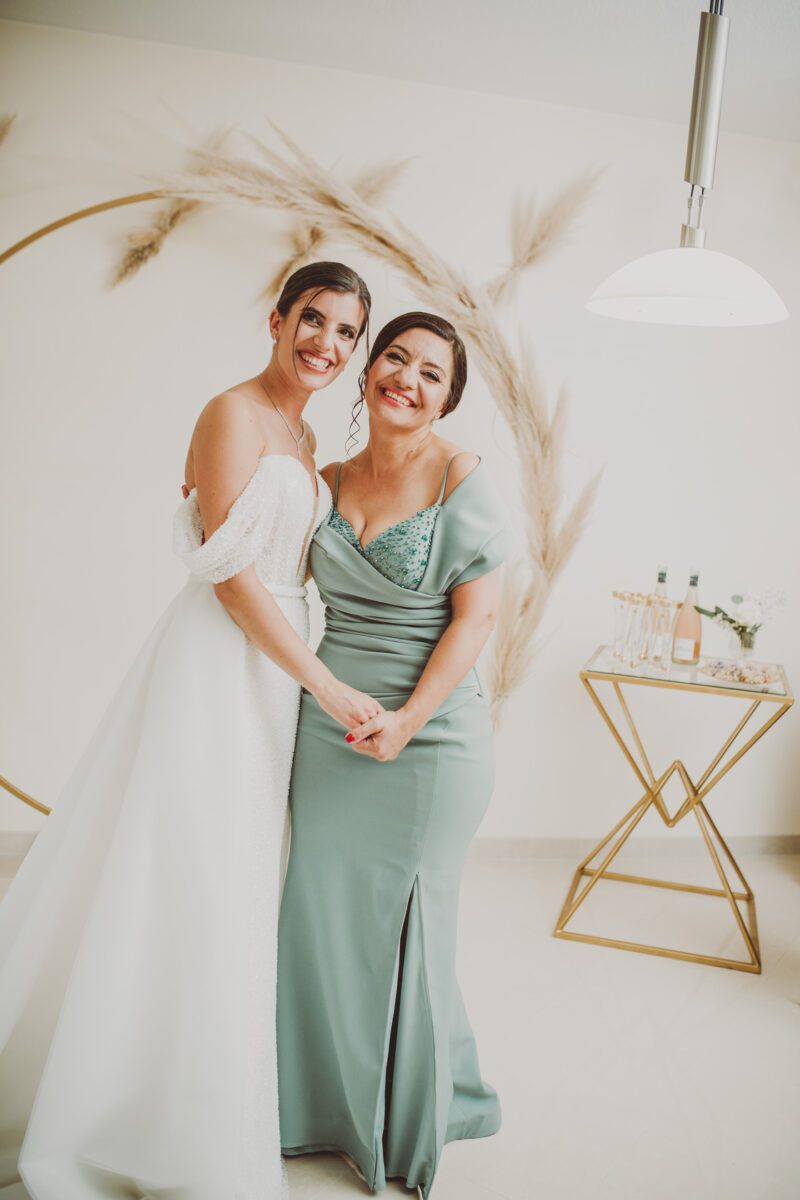 Zwei Frauen lächeln und halten sich an den Händen. Die eine trägt ein weißes, schulterfreies Brautkleid und die andere ein grünes V-Ausschnitt-Kleid mit Schlitz. Sie stehen vor einem runden Hintergrund, der mit Pampasgras geschmückt ist, wunderschön eingefangen von ihrem Frankfurter Hochzeitsfotografen. Auf der rechten Seite des Bildes steht ein kleiner Tisch mit Flaschen und Blumen. - Elis Wedding - Hochzeitsfotograf Frankfurt