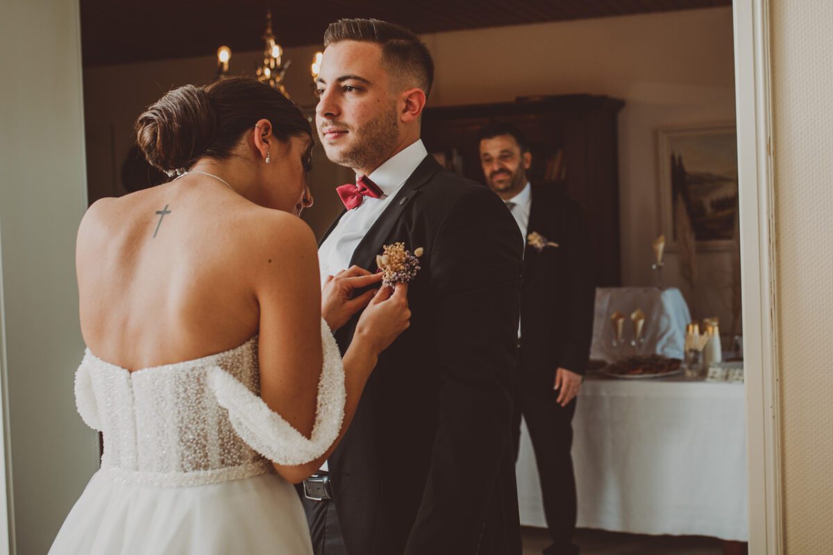Eine Braut, gekleidet in ein weißes trägerloses Brautkleid mit einem perlenartigen Mieder, steckt eine Boutonniere an das Revers des schwarzen Anzugs des Bräutigams. Der Bräutigam, der eine rote Fliege trägt, blickt nach vorne. Ein Mann im Anzug steht im Hintergrund, aufgenommen von ihrem Hochzeitsfotografen während der standesamtlichen Trauung. - Elis Wedding - Hochzeitsfotograf Frankfurt