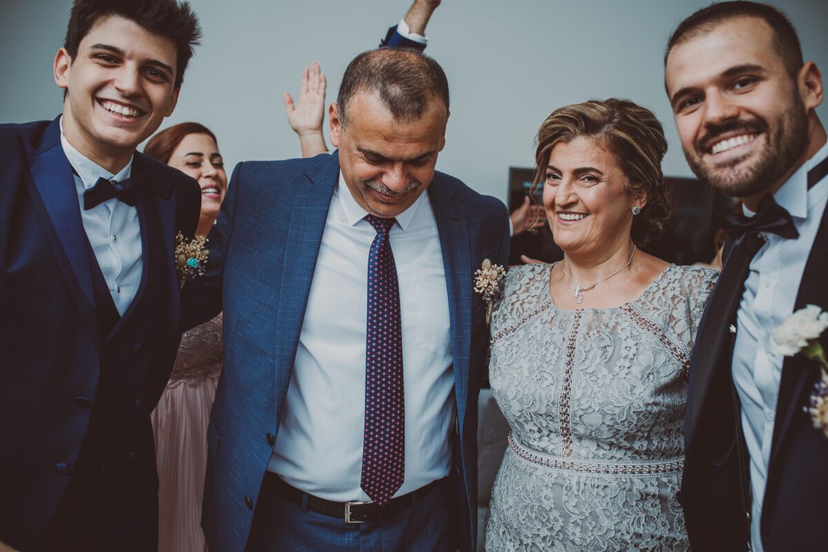 Eine Gruppe von fünf Personen lächelt und posiert gemeinsam. Der Mann in der Mitte, der einen blauen Anzug trägt, steht neben einer Frau in einem silbernen Spitzenkleid. Zwei Männer in Smokings flankieren sie auf beiden Seiten, und eine andere Person, die teilweise verdeckt ist, streckt im Hintergrund einen Arm aus. Die Kulisse lässt auf eine feierliche Veranstaltung in Frankfurt schließen, die von ihrem Hochzeitsfotografen festgehalten wurde. - Elis Wedding - Hochzeitsfotograf Frankfurt