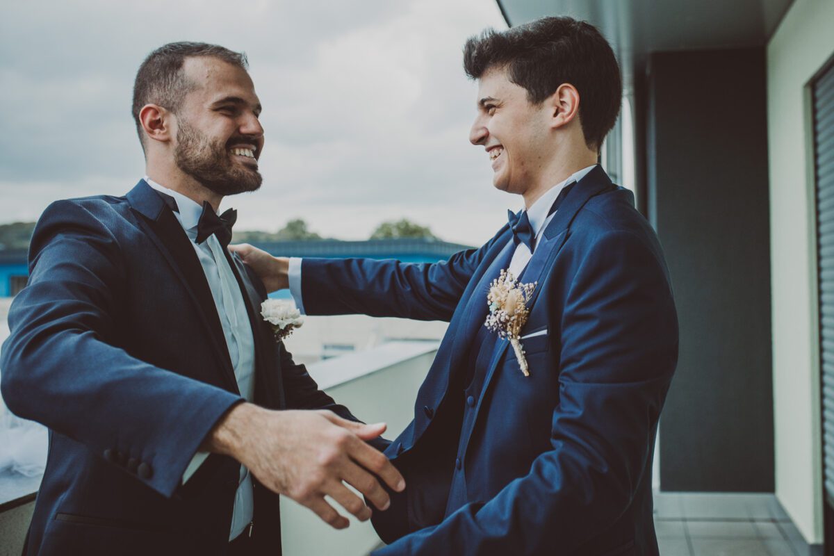 Zwei Männer in dunkelblauen Anzügen, weißen Hemden und schwarzen Fliegen lächeln und umarmen sich auf einem Balkon, wahrscheinlich genießen sie ein Fotoshooting. Beide tragen Boutonnieren; einer mit einer weißen Blume, der andere mit einem getrockneten Blumenarrangement. Der Hintergrund zeigt einen bewölkten Himmel und verschwommene Gebäude. - Elis Wedding - Hochzeitsfotograf Frankfurt