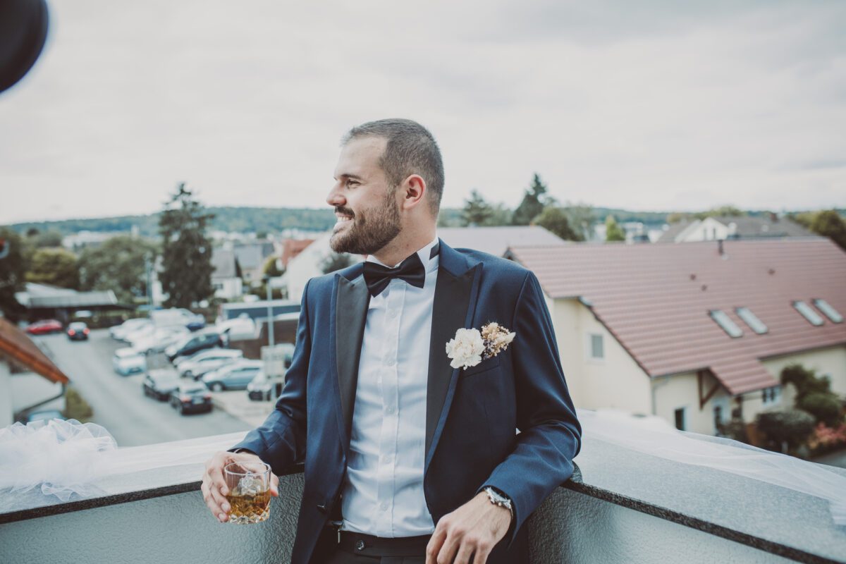Ein Mann mit gestutztem Bart und kurzen Haaren lehnt an einem Balkongeländer in Frankfurt und hält ein Getränk in der Hand. Er trägt einen dunklen Anzug, eine schwarze Fliege und ein weißes Oberhemd mit einer Blumenboutonnière an seiner Jacke und scheint bereit für ein Fotoshooting zu sein. Hinter ihm ist ein Blick auf Häuser und Grünflächen zu sehen. - Elis Wedding - Hochzeitsfotograf Frankfurt