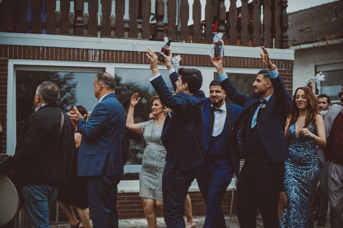 Eine lebhafte Feier im Freien, bei der mehrere Menschen energisch tanzen, klatschen und die Arme heben. Sowohl Männer als auch Frauen tragen formelle Kleidung, darunter Anzüge und elegante Kleider. Im Hintergrund ist ein Backsteingebäude mit weiß gerahmten Fenstern zu sehen; es fühlt sich an wie ein festliches Fotoshooting. - Elis Wedding - Hochzeitsfotograf Frankfurt