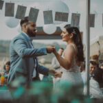 Braut und Bräutigam tanzen ihren ersten Tanz im Freien bei ihrer Hochzeitsfeier. Der Bräutigam trägt einen hellblauen Anzug und die Braut ein weißes Spitzenkleid. Gäste sitzen an Tischen mit Getränken und Blumen und beobachten sie. Oben hängen Papierlaternen und Fotos an einer Leine und verstärken die festliche Atmosphäre, die der Hochzeitsfotograf eingefangen hat. - Elis Wedding - Hochzeitsfotograf Frankfurt