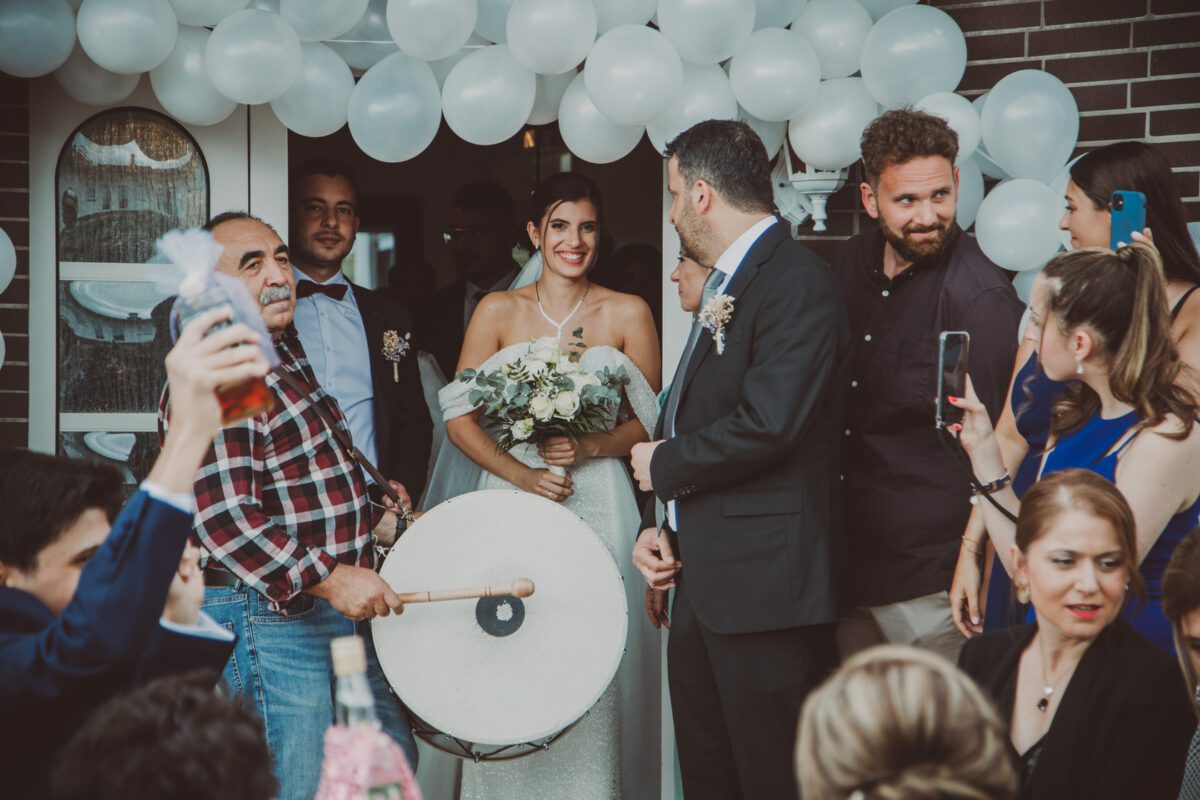 Eine lächelnde Braut in einem weißen Kleid hält einen Blumenstrauß in der Hand, während sie unter einem Ballonbogen am Eingang steht, umgeben von ihrer Hochzeitsgesellschaft. Der Bräutigam steht in der Nähe, den Gästen zugewandt. Ein Mann spielt eine Trommel, und die Gäste, in formeller Kleidung gekleidet, machen Fotos und feiern um sie herum – wunderschön eingefangen von unserem Hochzeitsfotografen aus Frankfurt. - Elis Wedding - Hochzeitsfotograf Frankfurt