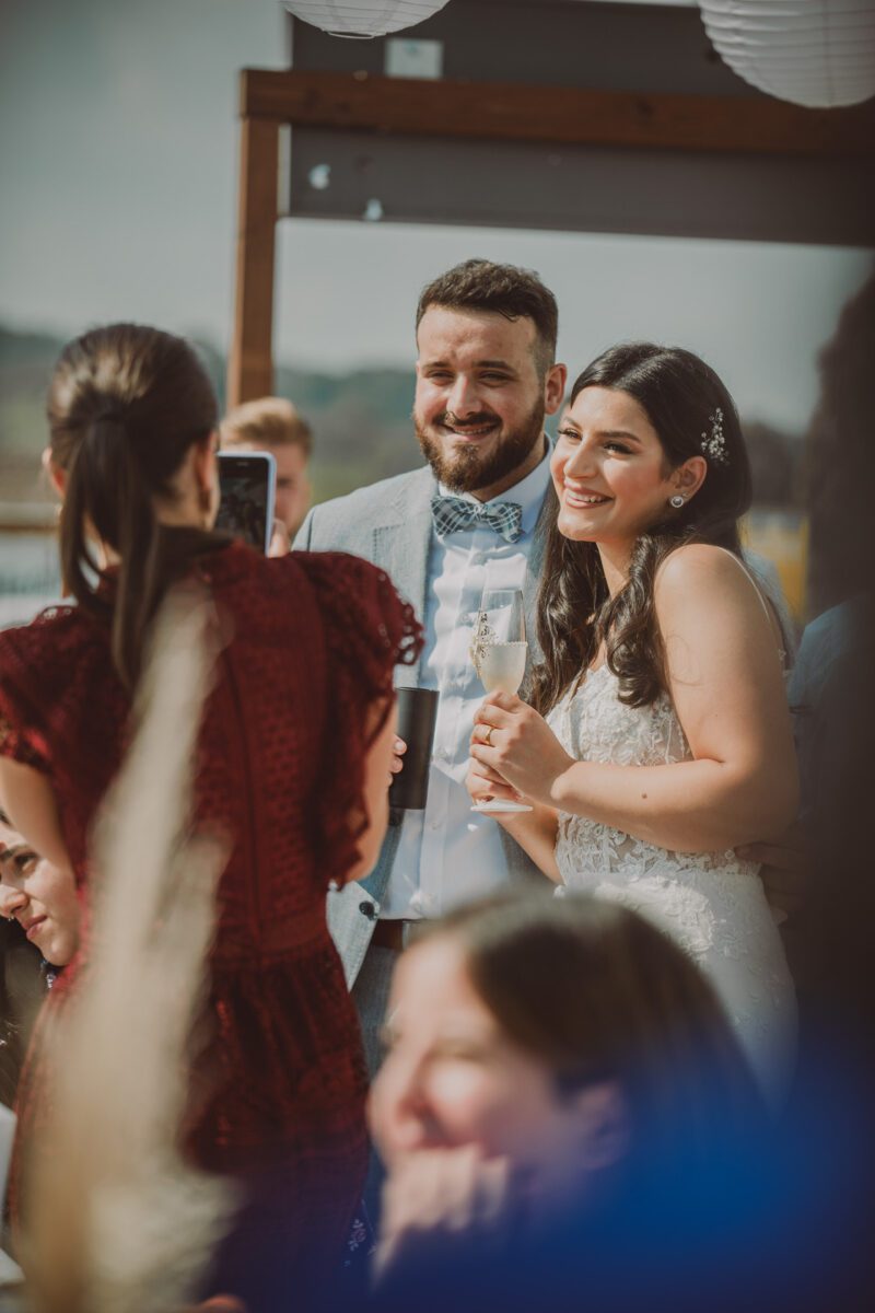 Braut und Bräutigam lächeln, während sie zu einem Gast blicken, der gerade mit einem Smartphone ein Foto macht. Die Braut in einem weißen Spitzenkleid hält ein Glas Champagner. Der Bräutigam in einem hellgrauen Anzug mit Fliege steht neben ihr. Diese Feier im Freien an einem sonnigen Tag fängt die Essenz der Fotografie ein. - Elis Wedding - Hochzeitsfotograf Frankfurt