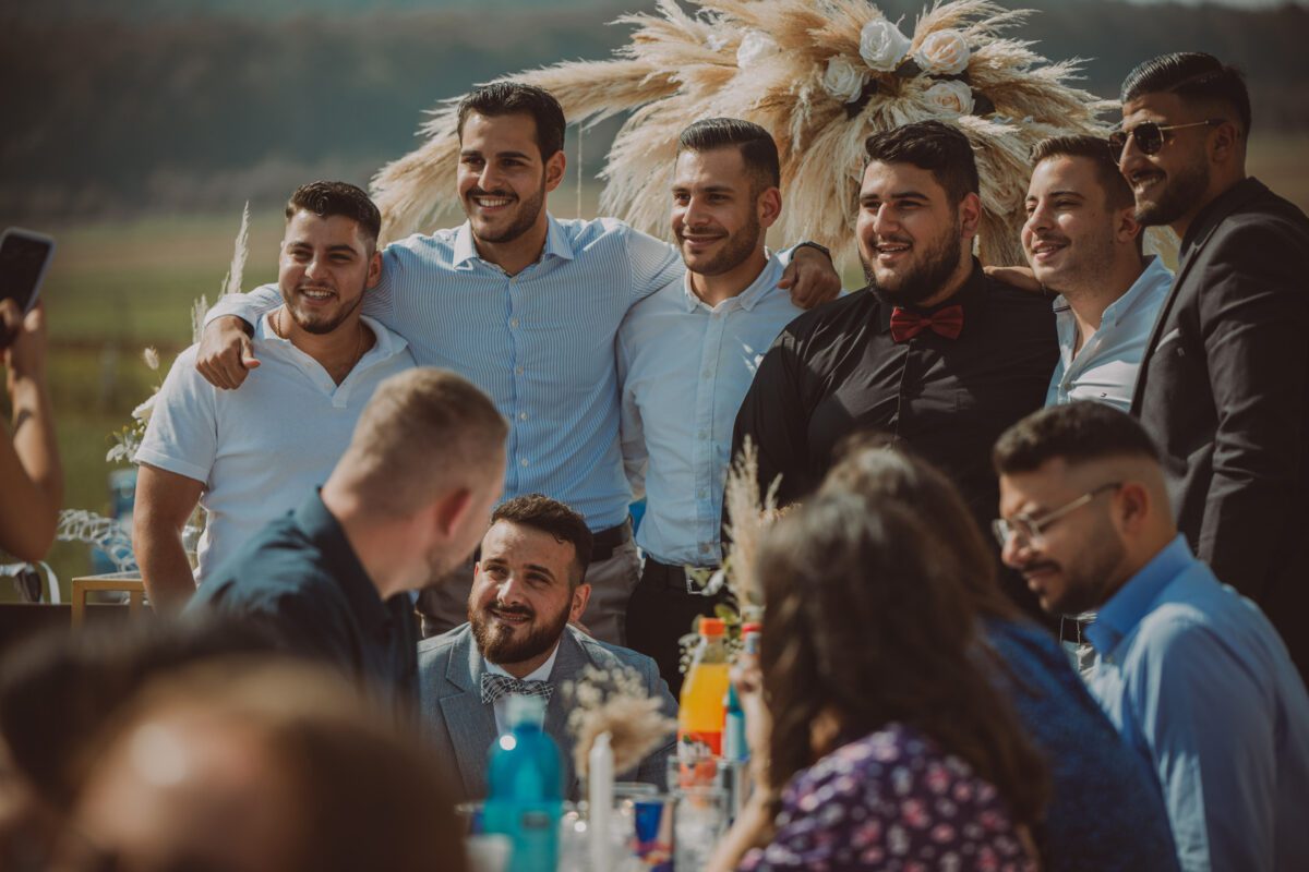 Eine Gruppe von sieben Männern steht zusammen im Freien, lächelt und posiert für ein Foto. Hinter ihnen ist ein großes Blumenarrangement mit hohen, federartigen Federn zu sehen. Im Vordergrund sitzen mehrere Personen an Banketttischen, die mit bunten Flaschen und Blumengestecken bedeckt sind. Die Fotografie fängt die hell erleuchtete Szene wunderschön ein. - Elis Wedding - Hochzeitsfotograf Frankfurt