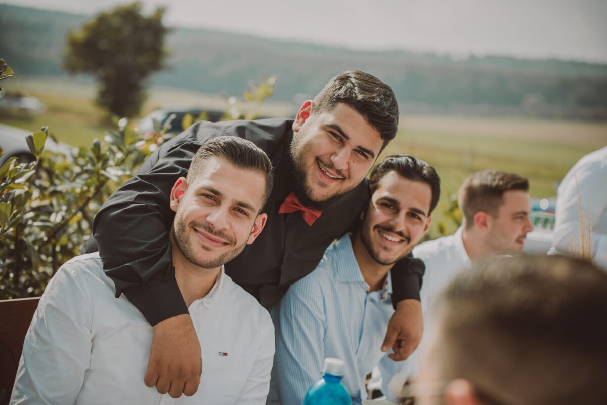Drei junge Männer lächeln in die Kamera. Einer in der Mitte trägt ein schwarzes Hemd und eine rote Fliege und legt seine Arme um seine Freunde in weißen Hemden. Sie sind draußen mit viel Grün im Hintergrund, zusammen mit verschwommenen weiteren Personen und malerischen Hügeln einer kürzlichen standesamtlichen Trauung. - Elis Wedding - Hochzeitsfotograf Frankfurt