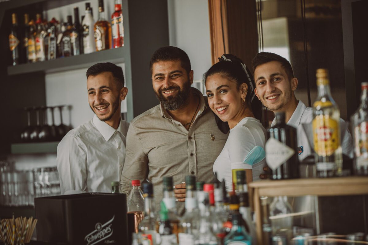 Eine Gruppe von vier Personen, zwei Männer und zwei Frauen, steht in Frankfurt hinter einer Bar, die mit verschiedenen Alkoholflaschen gefüllt ist. Sie lächeln alle in die Kamera. Im Hintergrund sind Regale mit weiteren Flaschen und Gläsern zu sehen, in der Mitte steht ein Mann mit Bart und einem hellen Hemd. - Elis Wedding - Hochzeitsfotograf Frankfurt