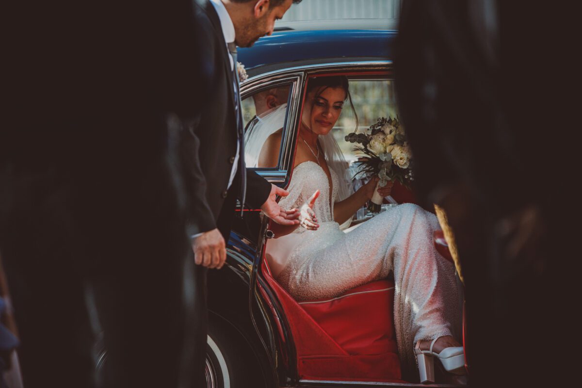 Eine Braut in einem strahlend weißen Kleid und Schleier, die einen Blumenstrauß in der Hand hält, lächelt, als sie aus einem schwarzen Auto mit roter Innenausstattung steigt. Ein Mann in einem dunklen Anzug reicht ihr seine Hand, um ihr zu helfen. Um sie herum sind teilweise andere Gestalten zu sehen, die die Atmosphäre dieses Frankfurter Hochzeitsfotoshootings einfangen. - Elis Wedding - Hochzeitsfotograf Frankfurt