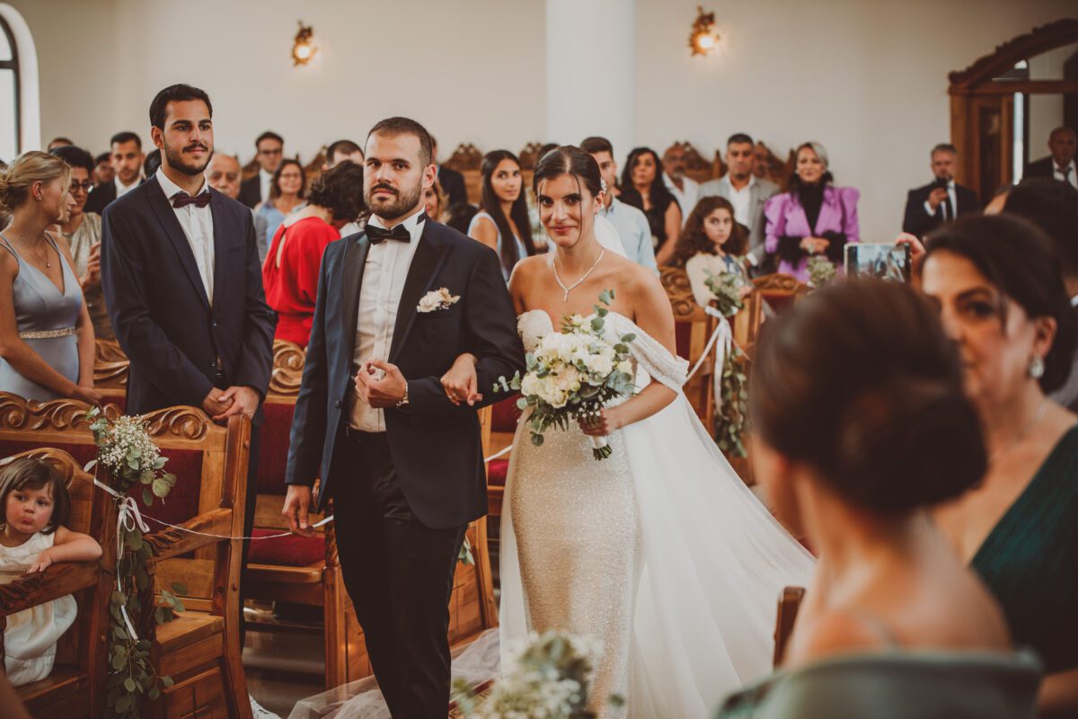 Eine Braut in einem schulterfreien weißen Kleid mit langem Schleier hält einen Blumenstrauß in der Hand und schreitet mit einem bärtigen Mann im Smoking den Gang entlang. Die elegant gekleideten Gäste sitzen und stehen und beobachten den Umzug in einem warm beleuchteten Saal mit kunstvollen Holzstühlen und Blumendekorationen – perfekt für jede Frankfurter Fotosession. - Elis Wedding - Hochzeitsfotograf Frankfurt